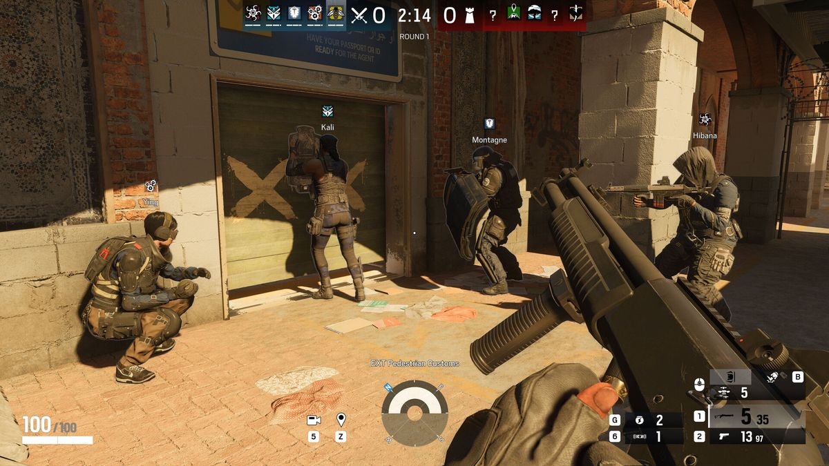 Capture officielle de Rainbow Six Siege : quatre attaquants se préparent devant un rideau métallique marqué de deux croix, l'une plaque un gadget contre la paroi pendant qu'un porteur de bouclier couvre l'entrée
