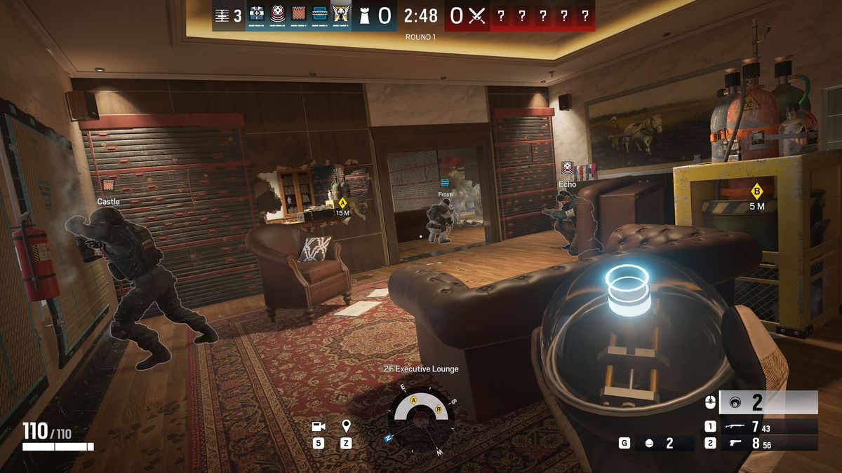 Capture officielle de Rainbow Six Siege : trois défenseurs préparent un salon à l'étage, un mur déjà percé laisse voir la pièce voisine, et le joueur tient un gadget prêt à être posé